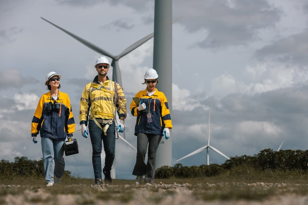 Three technicians in hard hats and hi-vis walking towards wind turbines, one carrying a tablet.