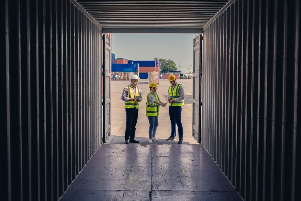 Three warehouse staff in hi-vis vests reviewing a tablet, framed by the open doors of a shipping container.