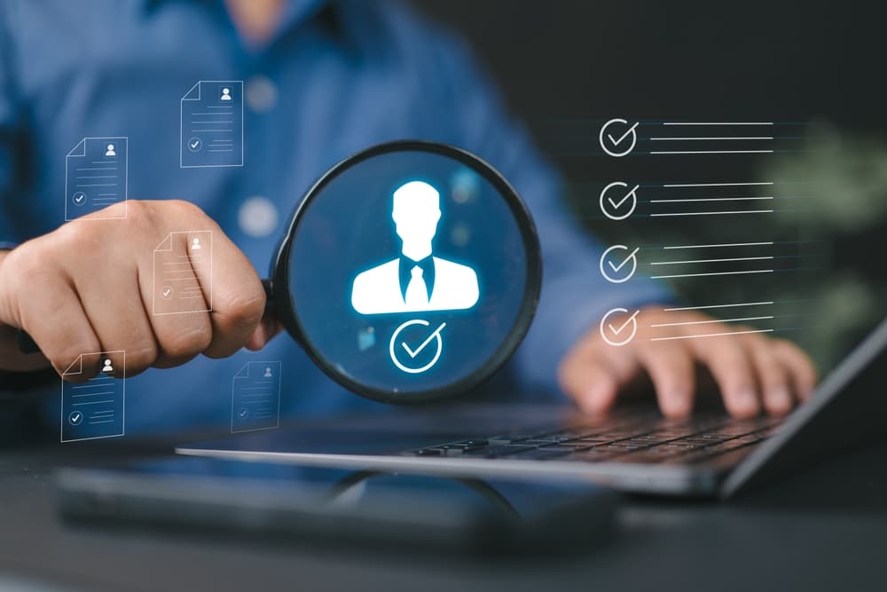 A recruiter's hand holding a magnifier over a glowing candidate profile above a laptop.